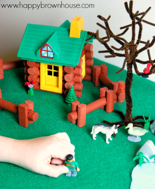 Child\'s hand playing with miniature toy person figurine. Log toy house, pipe cleaner tree, and animal toy figurines in the background. This children\'s activity is a Peter and the Wolf small world play activity for acting out the classical musical story.