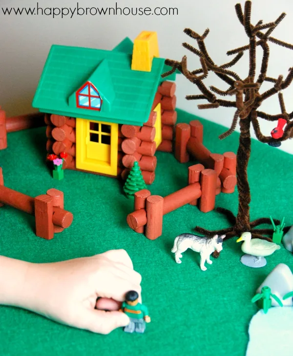 Child's hand playing with miniature boy toy figurine. Log toy house and animal toy figurines in the background. This children's activity is a Peter and the Wolf small world play activity for acting out the classical musical story.