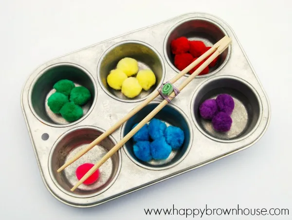 Metal muffin tin pan filled with colorful craft pom-poms. DIY Fine Motor Chopstick Tongs resting across the top of the muffin pan