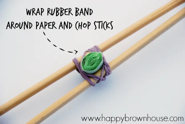 close up of how to attach the rubber band and 1 inch paper strip to the chopsticks to make the DIY Chopstick Fine Motor Tongs