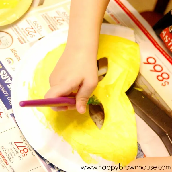 child\'s hand holding a purple paintbrush while painting a paper plate mask with yellow paint