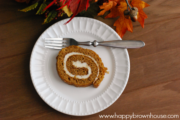 close up of a slice of spiraled pumpkin cake with cream cheese filling on a white plate with a fork, brown table and fall leaves in background