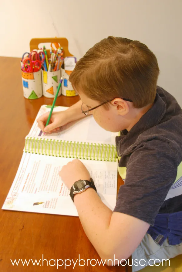 boy writing in a spiral workbook with a green pencil, colorful school supply caddy in the background