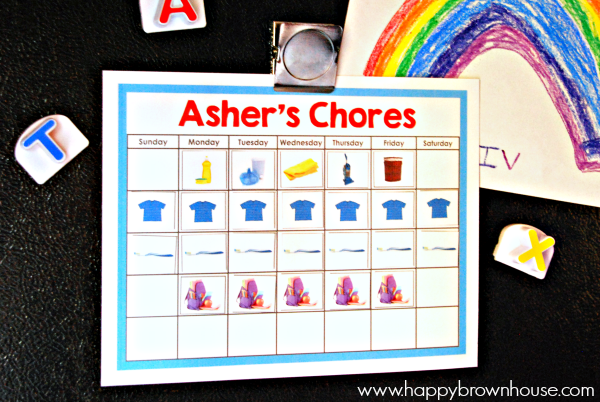 Editable Chore Chart hanging on a black refrigerator by a silver magnet surrounded by letter magnets and a child\'s rainbow artwork