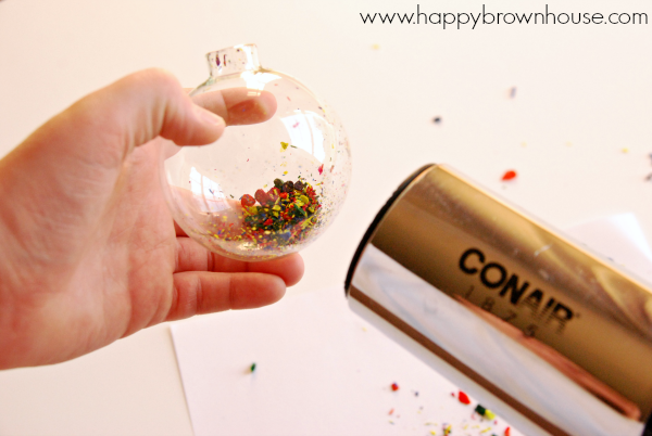 child\'s left hand holding a clear glass ornament with colorful crayon shavings inside, hair dryer being used to heat up the ornament and melt the crayon shavings