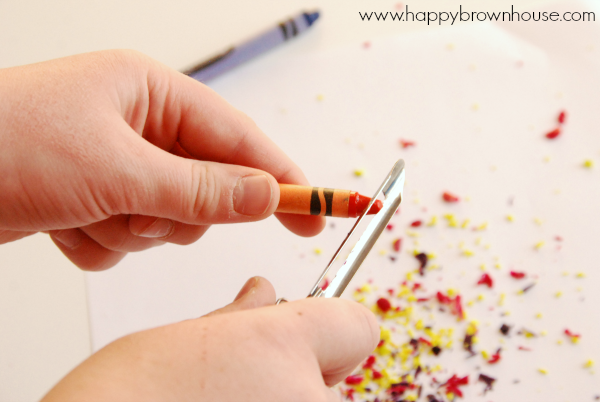 Child\'s hands holding an orange crayon in their left hand and a vegetable peeler in their right hand creating crayon shavings. Blue crayon and crayon shavings in the background on the white table