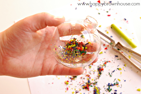 child\'s hand holding a clear glass ornament ball with the top removed, colorful crayon shavings have been placed inside the ornament