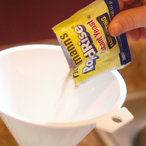 Child's hand pouring a yellow packet of instant yeast into a white funnel 