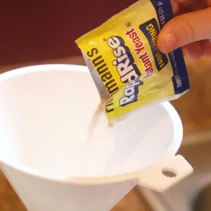 Child's hand pouring a yellow packet of instant yeast into a white funnel 