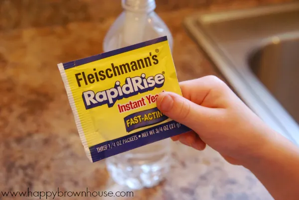 child\'s hand holding a yellow packet of Fleischmann\'s RapidRise instant yeast, clear plastic water bottle sitting on a brown speckled countertop in the background