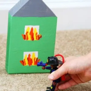 close up of child's hand playing pretend with PLAYMOBIL fire fighter figure and a wooden block house