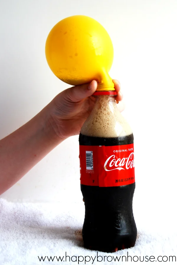 Close up of a child\'s hand holding a partially inflated yellow balloon attached to a Coca Cola bottle with fizz inside