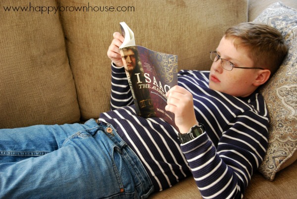 Boy in a blue and white striped shirt and jeans relaxing on a brown couch to read a book about Isaac Newton