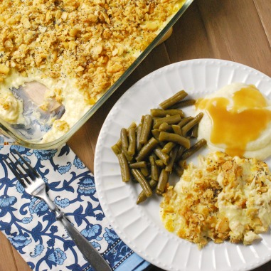 close up of chicken casserole, green beans, and mashed potatoes with gravy on a white plate beside a blue and white cloth napkin with a fork on top.