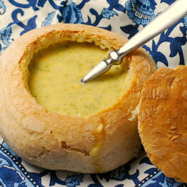 Homemade bread bowl holding Broccoli Cheese Soup and a silver spoon on a blue and white background.