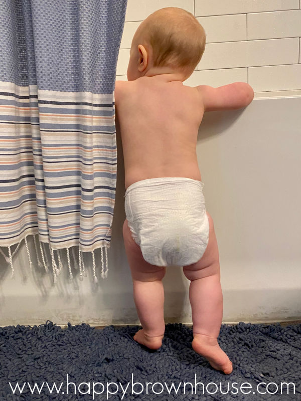 Cute baby in a diaper standing next to a bathtub waiting for the bathtub to fill with water