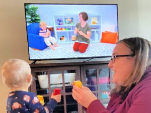 Mom and baby watching a television screen with another mom and baby on it. Mother and baby are holding colorful blocks to tap with the song rhythm