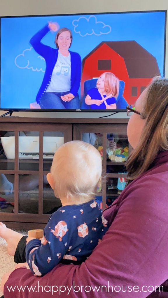 Mother holding a toddler sitting on her lap; both are watching an online music class on the television.