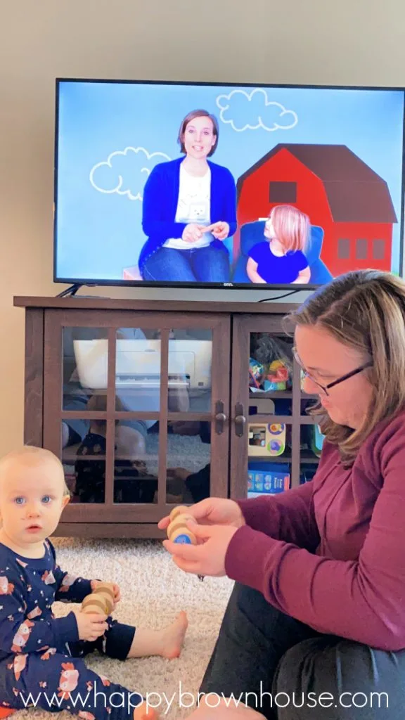 Mother and toddler holding wood toys and tapping them together to make rhythmic sounds. Television in the background has an online music class for the mother and baby.